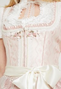 Femme portant une robe corset rose à motifs avec un galon en dentelle blanche, de petites décorations en roses roses et un grand nœud en satin blanc noué à la taille.