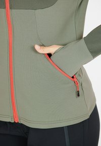 Olivengrüne Activewear-Jacke aus strukturiertem Stoff, ausgestattet mit einem leuchtend orangefarbenen Reißverschluss und einer Seitentasche, entworfen für Funktionalität und Komfort.