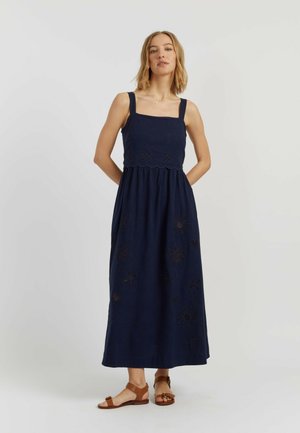 Femme debout portant une robe maxi bleu marine sans manches avec des motifs floraux découpés et des sandales plates marron sur un fond uni.