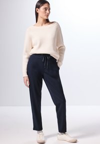Maglione a coste crema con scollo ampio abbinato a pantaloni jogger blu navy con cordino. Il modello indossa sneakers bianche, evidenziando il look informale.
