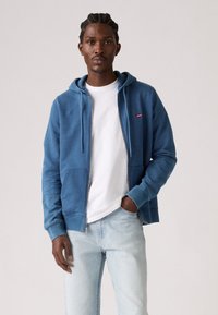 Blauwe rits-hoodie van zachte stof, met een voorkantzak en een capuchon met trekkoord. Gecombineerd met een wit T-shirt en lichtblauwe spijkerbroek.