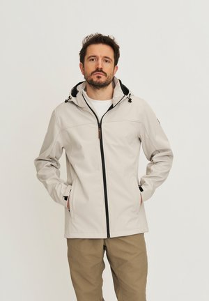 Man met een beige capuchonjas en kaki broek, staand met handen in de zakken tegen een effen achtergrond.