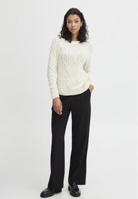 Crème gebreide trui met kabelpatroon, ronde hals en geribbelde mouwen, gecombineerd met zwarte wijde broek en zwarte schoenen.