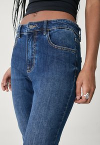 Dunkelblaue Jeans aus Denim mit einer figurschmeichelnden Passform, sichtbaren Nähten, Taschen und einem Knopfverschluss. Die Oberfläche hat eine glatte Textur.