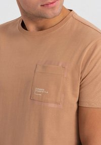 Lysebrun kortærmet t-shirt i blød bomuld med en tekstureret brystlomme, der har trykt logo. Rund hals og pæne stikninger.