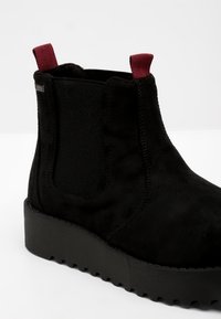 Bottine en daim noir avec une semelle épaisse noire crantée et des languettes rouges à l'avant et à l'arrière. Panneau latéral élastique pour un enfilage facile.