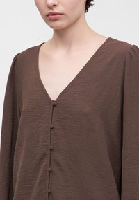 Jeune personne portant un chemisier marron texturé à col en V avec des boutons recouverts de tissu sur le devant et des manches longues.
