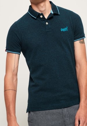 Mann trägt ein dunkelblaues Poloshirt mit hellblauen Abschlüssen und Logo, kombiniert mit grauer Hose. Sein Gesicht ist nicht sichtbar.