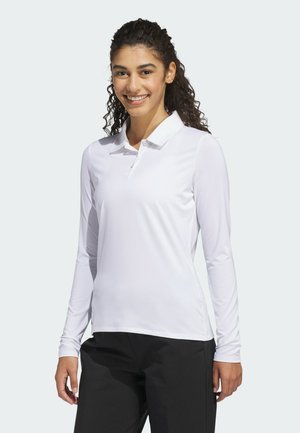 Ung kvinde med krøllet hår smiler, iført en hvid langærmet poloshirt og sorte bukser, stående mod en ensfarvet lys baggrund.