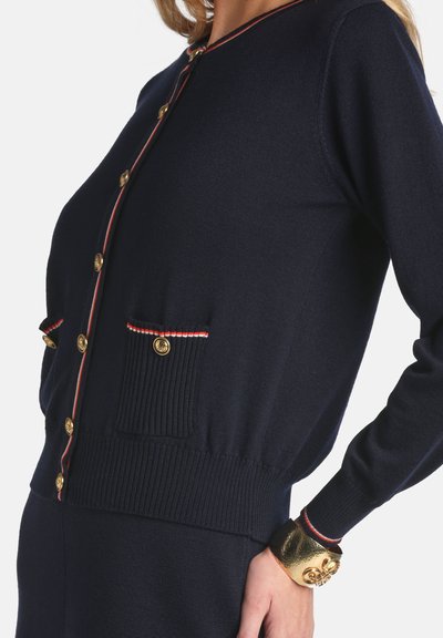 Marineblauer Strick-Cardigan mit goldenen Knöpfen und rotem Akzentbesatz. Verfügt über gerippte Bündchen und Taschen. Auffälliges goldfarbenes Designelement am Handgelenk.
