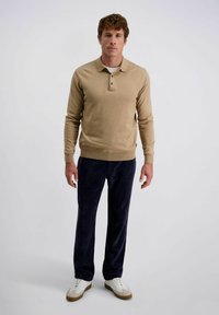 Man die staat en een beige lange mouwen polo, donkere blauwe corduroy broek en witte sneakers draagt, tegen een effen lichte achtergrond.