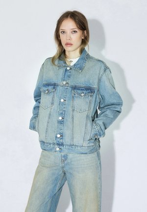 Młoda kobieta w jasnoniebieskiej dżinsowej kurtce i dżinsach, stoi z rękami w kieszeniach na jednolitym białym tle.