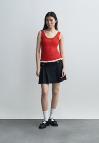 Νέα γυναίκα όρθια, φοράει κόκκινο αμάνικο τοπ με λευκή δαντέλα στη διακόσμηση, μαύρη πλισέ φούστα, λευκές κάλτσες και μαύρα παπούτσια.