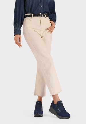 Jeans beige a vita alta con orlo sfilacciato abbinati a sneakers blu scuro senza lacci con cerniere e dettagli testurizzati. Una camicia in denim chiaro completa l'outfit.