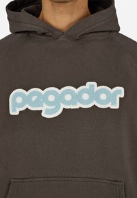 Barna kapucnis pulóver, amelyen egy nagy, világoskék hímzett logó található, melyen a "pegador" felirat olvasható vidám betűtípussal. Puha tapintású, elülső zsebbel.