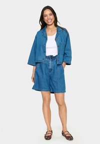 Denim jaka ar kvadrātveida piegriezumu un pogu aizdari, kombinēta ar augsto vidukļa denim šortiem. Abi ir vidēji zila krāsā, audums ar tekstūru.