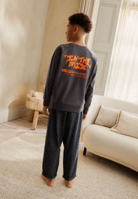 Grauer Pullover mit orangefarbenem "IN THE MIX" Schriftzug auf der Rückseite, kombiniert mit hoch taillierten, karierten Schlafanzughosen. Vor neutralem Hintergrund präsentiert.