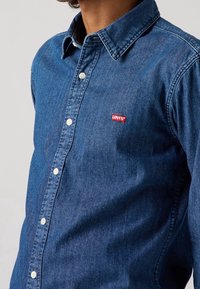 Donkerblauwe denim overhemd met een knoopsluiting, puntige kraag en een klein rood Levi's logo op de borst.