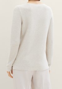 Maglione grigio chiaro a coste con maniche lunghe e dettaglio a chiave sulla schiena. Texture morbida e orlo leggermente curvato; indossato sopra pantaloni chiari.