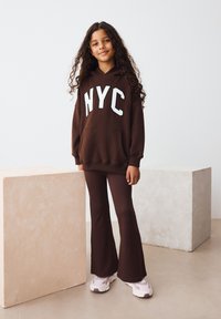 Sweat à capuche marron avec lettrage blanc "NYC", assorti à un pantalon marron évasé. Le mannequin porte des baskets rose pâle. Fond minimaliste.