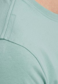 Camiseta de color teal claro con cuello redondo, mangas cortas y una textura suave y lisa, con detalles de costura visibles en el hombro.