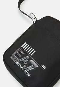 Borsetta in tessuto nero con finitura testurizzata, caratterizzata dal logo bianco "EA7 EMPORIO ARMANI" e da un design a righe, inclusa una tracolla regolabile.