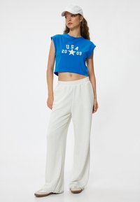 Camiseta azul corta con estampado de "USA 2009" y estrellas, combinada con pantalones anchos blancos y zapatos de lona beige. Una gorra blanca completa el look.