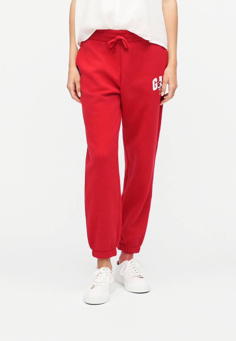 Άτομο που φοράει κόκκινη φόρμα με λευκό λογότυπο και κορδόνι στη μέση, συνδυασμένη με λευκά αθλητικά παπούτσια και χαλαρό λευκό μπλουζάκι.