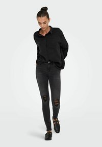 Camisa negra de botones con un bolsillo, combinada con unos jeans skinny rasgados negros y unos zapatos slip-on negros. Textura de tela suave.