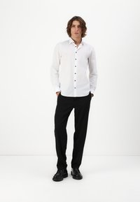 Weißes Hemd mit navyblauen Knöpfen, lange Ärmel, kombiniert mit schwarzen Hosen und schwarzen Schuhen, vor einem schlichten Hintergrund stehend.
