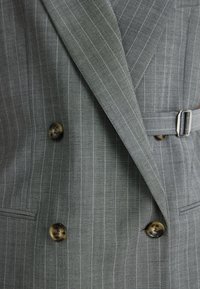 Harmaa raidallinen kaksirivinen bleiseri, jossa on hahlot, kaksi nappia ja hopeinen vyönsolkiyksityiskohta. Vaatteen kankaan rakenne on näkyvissä.