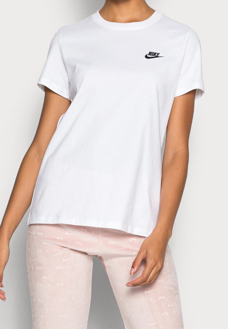 Kvinna iklädd vit Nike t-shirt och ljusrosa leggings med Nike Air-logotypmönster, stående mot en neutral bakgrund.
