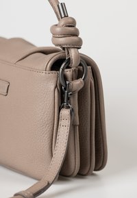 Un primo piano di una borsa in pelle taupe mostra una tracolla regolabile con nodo e un gancio in metallo nero attaccato agli anelli laterali.