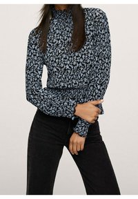 Blouse à manches longues présentant un motif floral noir et bleu clair, un col froncé et des poignets à volants. Associée à un jean noir.