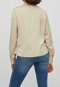 Femme portant un chemisier beige à manches longues et un jean bleu, debout de dos face à l'appareil photo sur un fond uni.