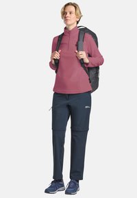 Maglione leggero in pile rosa con zip e maniche lunghe; pantaloni khaki navy con zip e tasche laterali; zaino grigio; scarpe da ginnastica blu.