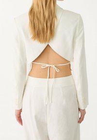 Blazer blanco cropped con un corte dramático en la espalda, atado a la cintura con un cordón delgado y terminado con acentos de botones en las mangas.