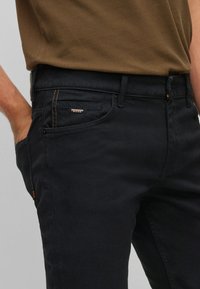 Mustat farkut slim fit -mallilla, joissa on oranssit ompeleet ja pieni logo-merkki. Kangas näyttää sileältä ja siinä on hieman kiiltoa.