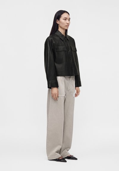 Femme portant une veste en cuir noire boutonnée, un pantalon beige large et des chaussures noires pointues, debout devant un fond blanc uni.