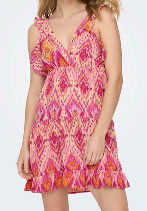 Vrouw die een mouwloze jurk met V-hals draagt, met een patroon van roze, oranje en paarse ruiten, en met ruches aan de zoom en schouderbandjes.