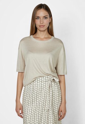 Woman with straight brown hair wears beige short-sleeve top and white wrap skirt with black square pattern, standing against white background.