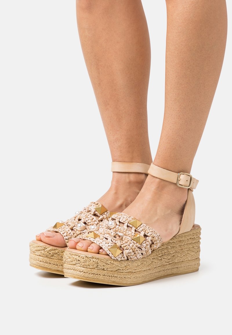 Pieds chaussés de sandales à plateforme beiges tissées avec des brides autour de la cheville, décorées de clous dorés et de perles, sur un fond clair uni.