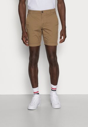 Shorts en coton marron avec une fermeture à bouton, un ourlet retroussé et deux poches latérales. Associés à des baskets blanches et des chaussettes blanches avec des rayures rouges et bleues.