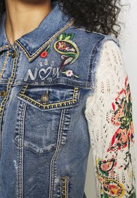 Džínová bunda s vyšívanými květinovými vzory, metalickými knoflíky a krajkovým designem rukávů. Modrá barva se zdobným prošitím.