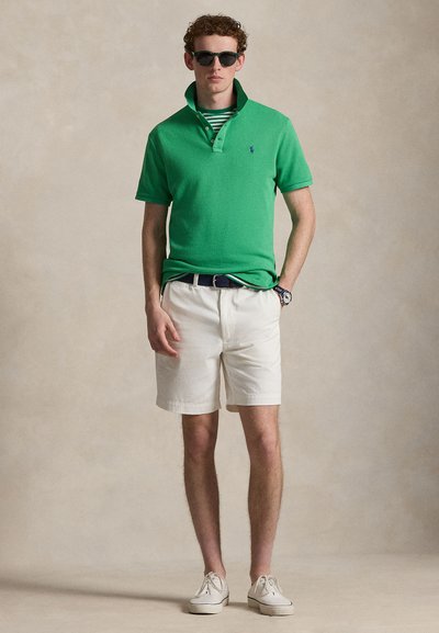 Νέος άνδρας που φοράει πράσινο πόλο πουκάμισο, λευκό σόρτς, σκουρόχρωμα γυαλιά ηλίου, λευκά αθλητικά παπούτσια και ρολόι χειρός, στέκεται με το ένα χέρι στην τσέπη.