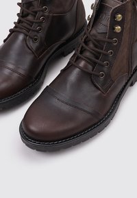 Bottines en cuir marron à lacets, avec un empiècement en daim contrastant, bout rond et semelle en caoutchouc texturée. Elles présentent des œillets en métal et des détails de couture.