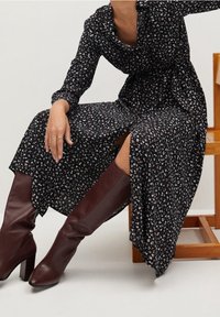 Lungo abito nero floreale con design avvolgente, maniche lunghe e spacco laterale. Abbinato a stivali in pelle bordeaux fino al ginocchio.