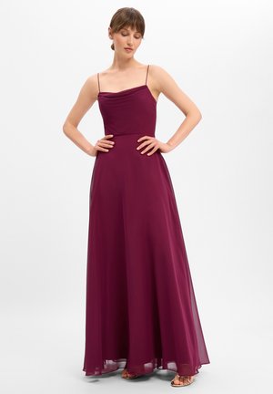 Frau trägt ein ärmelloses, bodenlanges, dunkelviolettes Kleid mit dünnen Trägern und steht mit den Händen in der Hüfte vor einem einfarbigen Hintergrund.
