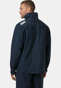 Chaqueta impermeable de color navy con alto collar, puños ajustables y logo blanco en el hombro. Presenta un diseño elegante y funcional con tejido texturizado.