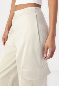 Pantaloni cargo beige chiaro in un tessuto morbido e texturizzato, dotati di una tasca laterale, logo ricamato e chiusura con zip in vita.